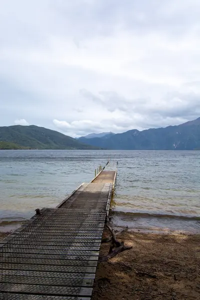 Sakin bir göle uzanan, bulutlu bir gökyüzünün altında dağlarla çevrili sakin bir ahşap iskele. Su manzarayı yansıtır, sakin bir atmosfer yaratır..