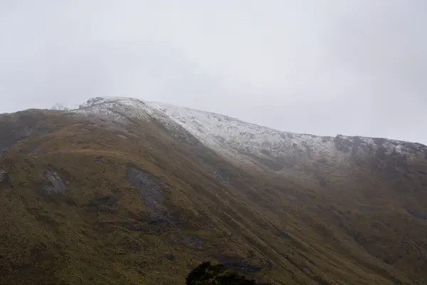 Karla kaplı tepe ve engebeli arazi içeren sisli bir dağ manzarası. Sahne bulutlu gökyüzü ve yeşil ve kahverengi bitki örtüsü karışımı ile karakterize edilir..