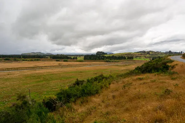 Bulutlu bir gökyüzünün altında çimenli bir tarlanın yer aldığı engin bir manzara. Sahnede uzak tepeler ve kıvrımlı bir yol, yeşil alanlar ve ön planda kuru otlar yer alıyor..