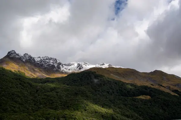 Bulutlu bir gökyüzünün altında yemyeşil ormanlarla çevrili karla kaplı dağların nefes kesici manzarası. Kara bulutlar ve parlak tepeler arasındaki zıtlık dramatik bir manzara yaratır..