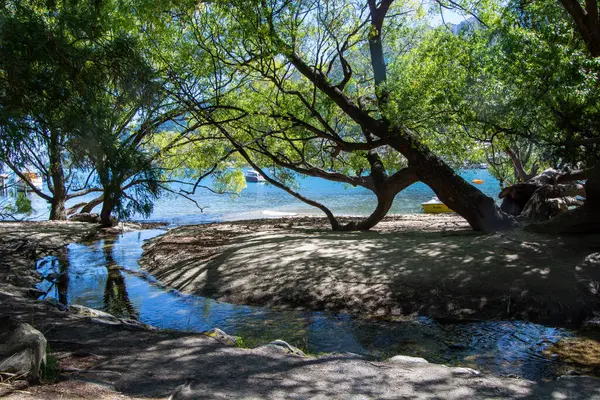 Bereketli yeşil ağaçlarla çevrili, kumlu bir alandan akan sakin bir su akıntısının olduğu sakin bir manzara. Güneş ışığı yaprakların arasından süzülerek yerde benekli bir ışık oluşturur. Arka planda, sakin bir göl görülebilir, tekneleri G