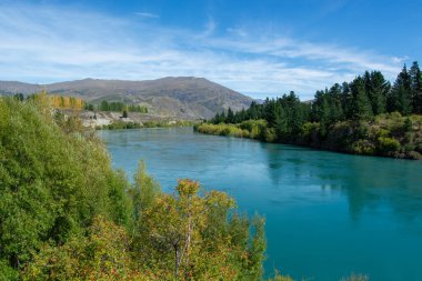 Berrak bir mavi gökyüzünün altında, ağaçlar ve dağlarla çevrili, sakin bir nehir, yemyeşil bir arazide akar. Su, çevrenin doğal güzelliğini yansıtan canlı bir turkuazdır..