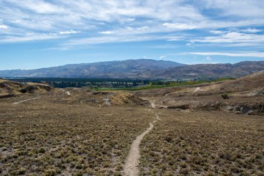 Kuru çimenler ve seyrek bitkiler arasında uzanan dolambaçlı bir toprak yolu barındıran uçsuz bucaksız, kurak bir arazi. Arka planda, yuvarlanan tepeler ve dağlar dağınık bulutlarla mavi bir gökyüzünün altında görülebilir..