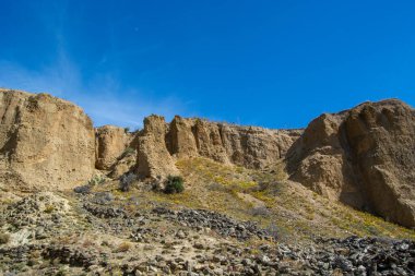 Parlak mavi bir gökyüzünün altında dik ve kumlu uçurumlar bulunan engebeli bir manzara. Arazi bitki örtüsü ve dağınık kayalarla kurumuş, doğal ve kurak bir ortam sergiliyor..