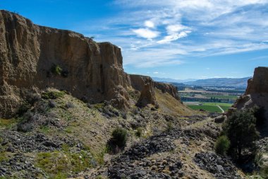 Engebeli uçurumların manzarası ve dağınık bulutlu açık mavi gökyüzünün altındaki kayalık arazi. Manzara yeşil alanlar ve uzak dağları barındırıyor, sakin bir doğal ortam yaratıyor..