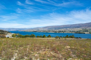 Yuvarlanan tepeler ve berrak mavi gökyüzü ile çevrili sakin bir göl manzarası. Ön planda seyrek bitki örtüsüne sahip çimenli bir alan yer alırken, arka planda dağlar ve su kenarında dolambaçlı bir yol yer alıyor..