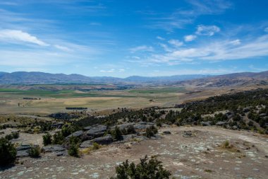 Uçsuz bucaksız bir manzaranın panoramik bir görüntüsü. Uçsuz bucaksız tepeler, tarlalar ve açık mavi gökyüzünün altında dağınık bulutlu uzak dağlar. Ön planda kayalık arazi ve seyrek bitki örtüsü yer alıyor..