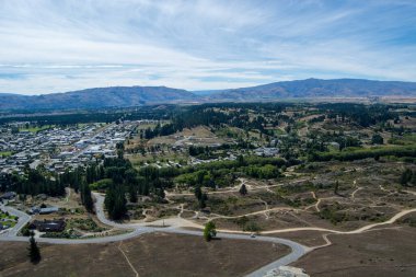 Tepeler ve yeşilliklerle çevrili, dolambaçlı yollar ve açık arazilerle çevrili küçük bir kasabanın havadan görünüşü. Manzara, açık mavi bir gökyüzü altında yerleşim alanları ve doğal manzaraların bir karışımını içerir..