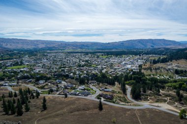 Dağlar ve yeşilliklerle çevrili küçük bir kasabanın havadan görünüşü, üzerinde açık mavi gökyüzü olan evler ve yollardan oluşan bir şebekeye sahip..
