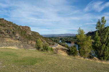 Bereketli yeşilliklerle çevrili dolambaçlı bir nehrin manzarası ve mavi gökyüzünün altında yuvarlanan tepeler ince bulutlarla. Ön planda nehir kenarına uzanan çimenli arazi yer alıyor..