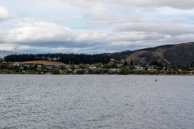 Tepeler ve küçük bir kasabayla çevrili bir göl manzarası. Gökyüzü parçalı bulutlu, sakin bir atmosferi yansıtıyor. Ön planda su bulunurken, arka planda tepedeki konut binaları ve yeşillikler sergileniyor..