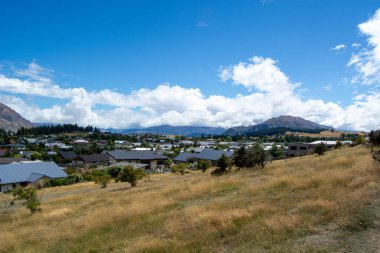 Dağlar ve mavi gökyüzüyle çevrili küçük bir kasabanın manzarası. Ön planda kuru çimenler ve çalılar yer alırken, arka planda ise evler ve parçalı bulutlu bir gökyüzünün altındaki uzak tepeler yer alıyor..