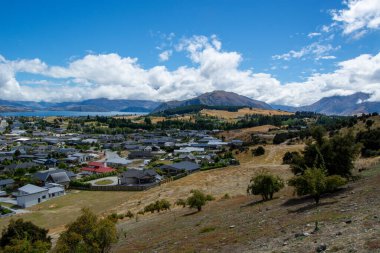 Dağlarla çevrili bir vadide küçük bir kasabanın manzarası. Evlerin ve yeşil manzaraların karışımı. Parlak mavi gökyüzü altında tüylü bulutlar..