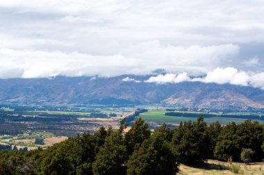 Bulutlu bir gökyüzünün altında yuvarlanan tepelerin ve dağların manzarası. Ön planda yemyeşil ağaçlar yer alırken, arka planda tarlalar ve uzak dağlarla dramatik bir manzara sergileniyor..