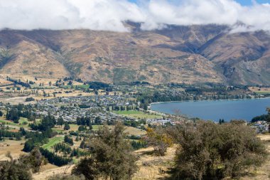 Dağlar, bir göl ve vadide yuva yapmış küçük bir kasabanın yer aldığı panoramik bir manzara. Sahne yuvarlanan tepeler, yemyeşil yemyeşil ve parçalı bulutlu gökyüzü ile karakterize edilir..