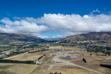 Dağların, tarlaların ve dağınık bulutlarla dolu açık mavi gökyüzünün bulunduğu engin bir manzaranın havadan görüntüsü. Olay yerinde tarım arazisi ve uzak bir inşaat sahası sergileniyor.