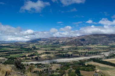 Parçalı bulutlu bir gökyüzünün altındaki dağlarla çevrili yemyeşil bir vadinin panoramik görüntüsü. Manzara, doğanın güzelliğini gösteren tarlalar, ağaçlar ve bir nehir yatağı içerir..
