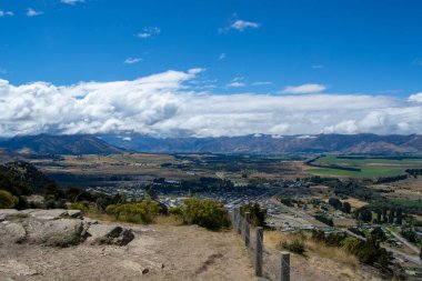 Parçalı bulutlu bir gökyüzünün altındaki dağlarla çevrili bir vadinin panoramik görüntüsü. Ön planda kayalık arazi ve çalılar yer alırken, arka planda verimli alanlar ve uzak tepeler yer alıyor..