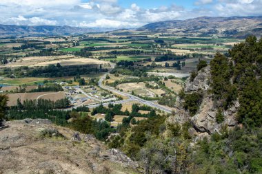 Dağlarla çevrili yemyeşil bir vadinin panoramik manzarası. Tarlalar, ağaçlar ve dolambaçlı bir yol. Gökyüzü parçalı bulutlu, sakin bir manzara yaratıyor..