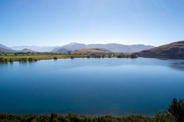 Bereketli yeşil tarlalar ve açık mavi gökyüzünün altındaki uzak dağlarla çevrili sakin bir göl. Su sakin, manzarayı yansıtıyor, huzurlu ve sakin bir atmosfer yaratıyor..