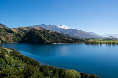 Berrak bir mavi gökyüzünün altında yemyeşil ve dağlarla çevrili sakin bir göl. Su, manzarayı yansıtır ve sakin bir manzara yaratır..