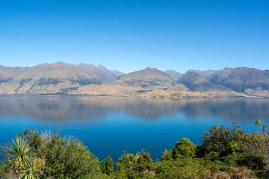Berrak mavi gökyüzünün altındaki dağları yansıtan sakin bir göl içeren sakin bir manzara. Yeşillik ön planda huzur sahnesine derinlik katıyor..