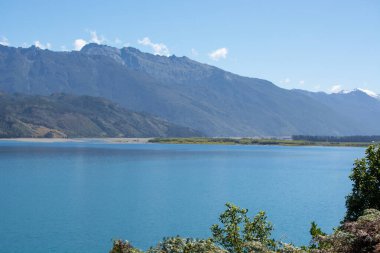 Berrak mavi gökyüzünün altında dağlarla çevrili sakin bir göl içeren sakin bir manzara. Su, doğanın canlı renklerini yansıtıyor ve barışçıl bir atmosfer yaratıyor..