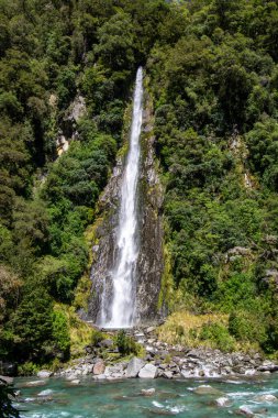 Büyüleyici bir şelale, yemyeşil bitki ve ağaçlarla çevrili kayalık bir uçurumdan aşağı yuvarlanıyor. Su, aşağıdaki turkuaz bir nehre akar ve sakin bir doğal manzara yaratır..