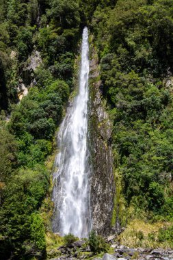 Sersemletici bir şelale, yemyeşil bitki örtüsüyle çevrili kayalık bir uçurumdan aşağı yuvarlanıyor. Su güçlü bir şekilde akıyor, tabanda bir sis yaratıyor, manzaranın doğal güzelliğini vurguluyor..