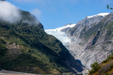 Gökyüzünde açık mavi gökyüzü ve ön planda yemyeşil yemyeşil yemyeşil, yüksek dağların arasına yerleşmiş bir buzulun çarpıcı görüntüsü. Buzul güneş ışığında parlıyor, buz mavisi renklerini sergiliyor..