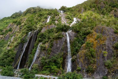 Etrafı yemyeşil bitki örtüsü ve sisli bulutlarla çevrili kayalık bir uçurumdan dökülen birden fazla şelalenin çarpıcı görüntüsü. Sahne, huzurlu bir ortamda doğanın güzelliğini yakalar..