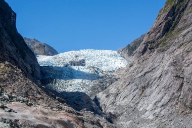 Berrak mavi gökyüzünün altında sarp kayalıklar arasında yuva yapmış bir buzulun nefes kesici görüntüsü. Buzulun buzlu yüzeyi, engebeli araziyle çelişerek gün ışığında parlıyor..