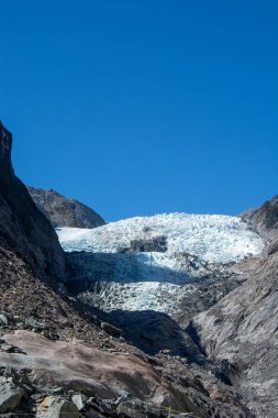 Berrak mavi gökyüzünün altındaki kayalık dağların arasına yerleştirilmiş bir buzulun çarpıcı görüntüsü. Buzul beyaz ve mavinin tonlarını sergiliyor. Çevresindeki arazinin toprak tonlarıyla çelişiyor..