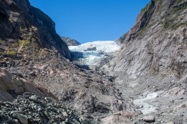 Berrak mavi gökyüzünün altında yükselen kayalık dağların arasına yerleşmiş bir buzulun çarpıcı görüntüsü. Buzulun buzlu yüzeyi engebeli araziyle çelişiyor, doğanın güzelliğini gözler önüne seriyor..