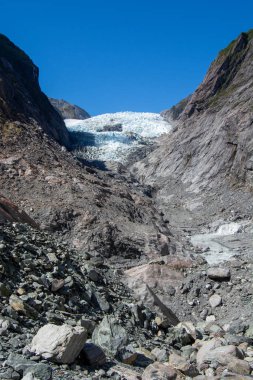 Berrak mavi gökyüzünün altındaki kayalık dağların arasına yerleştirilmiş bir buzulun çarpıcı görüntüsü. Buzul kısmen toprak ve kayalarla kaplı, engebeli araziyi gözler önüne seriyor..