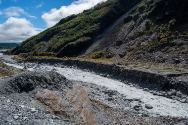 Pofuduk bulutlu, parlak mavi gökyüzünün altında yemyeşil tepelerle çevrili kayalık bir nehir yatağı. Nehir berrak suyla sığ ve manzara erozyon belirtileri gösteriyor..