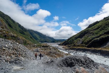 Bereketli yeşil dağlarla çevrili manzaralı bir vadide, kayalık bir nehir yatağı boyunca yürüyen iki yürüyüşçü parlak mavi bir gökyüzünün altında, tüylü bulutlarla..