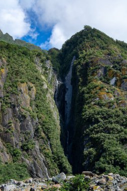 Yeşilliklerle kaplı sarp bir dağın heybetli manzarası. Kayalık uçurumdan şelale düşüyor. Gökyüzü parçalı bulutludur, manzaraya derinlik katar..