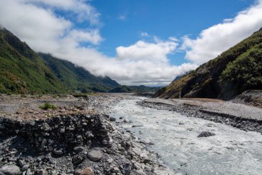 Parçalı bulutlu bir gökyüzünün altındaki dağlarla çevrili kayalık bir arazide akan bir nehrin manzarası. Nehir berrak ve mavi gökyüzünü yansıtırken yemyeşil kıyılar yemyeşil..