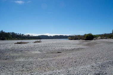 Açık mavi bir gökyüzünün altında, çakıl taşları ve seyrek bitki örtüsü ile kurumuş bir nehir yatağını barındıran huzurlu bir manzara. Sahne doğanın huzurunu yakalıyor, ufukta süzülen uzak ağaçlarla..