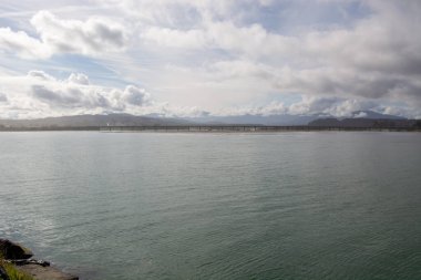 Sakin bir su kütlesi, uzak bir köprü ve bulutlu bir gökyüzünün altındaki dağlar. Sahne, suyun yüzeyine yansıyan yumuşak ışığı yansıtan doğanın sükunetini yakalar..