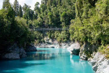 Bereketli yeşil ağaçlar ve arka planda ahşap bir asma köprü ile çevrili kayalık bankalar arasında akan turkuaz bir nehrin yer aldığı huzurlu bir manzara..