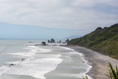 Dalgaların kıyıya vurduğu engebeli bir kıyı şeridi manzarası, yemyeşil tepeler ve bulutlu bir gökyüzünün altında kayalık oluşumlarla çevrili..