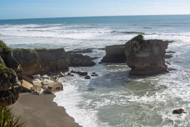 Kayalık oluşumlar ve dalgaların sahile çarptığı manzaralı bir kıyı manzarası. Gökyüzü birkaç bulutla berrak ve okyanus sakin, sakin bir atmosfer yaratıyor..