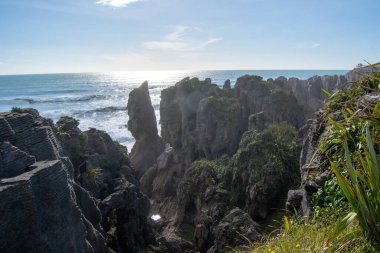 Engebeli kaya oluşumlarının ve açık mavi gökyüzü altında dalgaların çarpıştığı manzaralı bir kıyı manzarası. Güneş ışığı okyanustan yansıyarak sakin bir atmosfer yaratır..