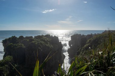 Engebeli uçurumların ve yemyeşil yeşilliklerin yer aldığı manzaralı bir sahil manzarası. Güneş okyanusun üzerinde parlıyor ve suyun yüzeyinde parıltılı bir etki yaratıyor..