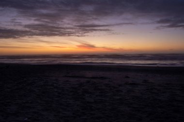 Gün batımında sakin bir sahil sahnesi, yumuşak dalgalar kıyıya vuruyor. Gökyüzü turuncu, pembe ve mor renklerle boyanarak sakin bir atmosfer yaratıyor. Kumsalın büyük bölümü boş, rahatlatıcı ve rahatlatıcı..