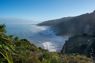 Bereketli yeşillik ve açık mavi gökyüzü ile çevrili kayalık kayalıklara çarpan dalgaların olduğu manzaralı bir kıyı manzarası. Güneş ışığı, manzaranın üzerine sıcak bir ışık saçıyor ve sakin bir atmosfer yaratıyor..