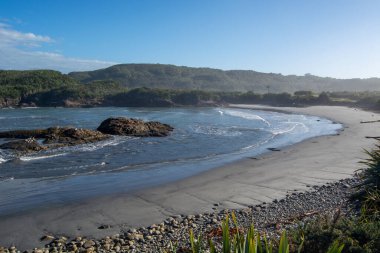 Sahilde hafif dalgaların, suda kayalık oluşumların ve açık mavi gökyüzünün altında yemyeşil tepelerin olduğu sakin bir sahil sahnesi..