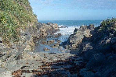 Sahil boyunca kayalık oluşumlar, yumuşak dalgalar ve yemyeşil suların yer aldığı sakin bir sahil manzarası. Gökyüzü parçalı bulutlu, sakin bir atmosfer yaratıyor..